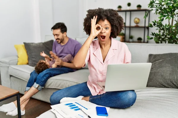 Mother of interracial family working using computer laptop at home doing ok gesture shocked with surprised face, eye looking through fingers. unbelieving expression.