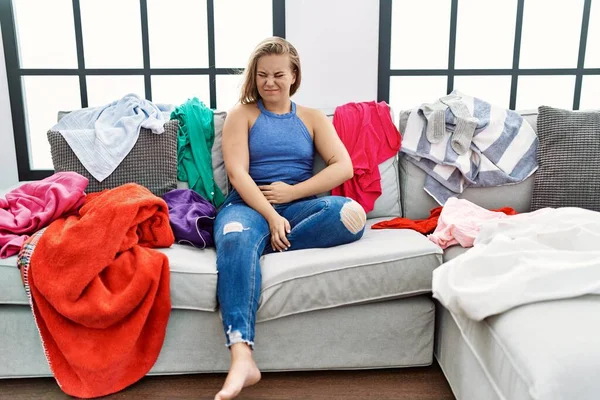 Young caucasian woman sitting on the sofa at home around dirty laundry with hand on stomach because indigestion, painful illness feeling unwell. ache concept.