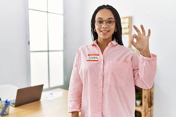 Young african american woman wearing hello my name is sticker identification doing ok sign with fingers, smiling friendly gesturing excellent symbol