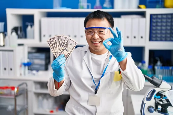Young chinese man working at scientist laboratory holding money doing ok sign with fingers, smiling friendly gesturing excellent symbol