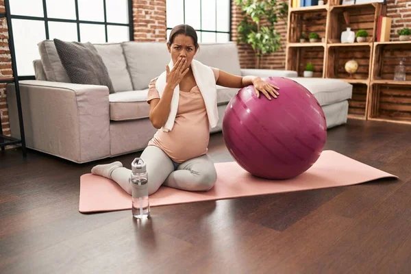 年轻的孕妇坐在瑜伽垫上 手拿着普拉提球捂着嘴 很震惊 很害怕犯错 惊讶的表情 — 图库照片