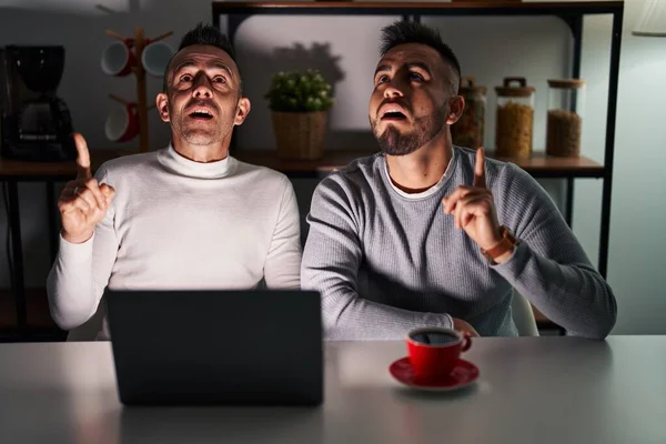 Homosexual couple using computer laptop amazed and surprised looking up and pointing with fingers and raised arms.
