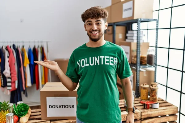 Young arab man wearing volunteer t shirt at donations stand smiling cheerful presenting and pointing with palm of hand looking at the camera.