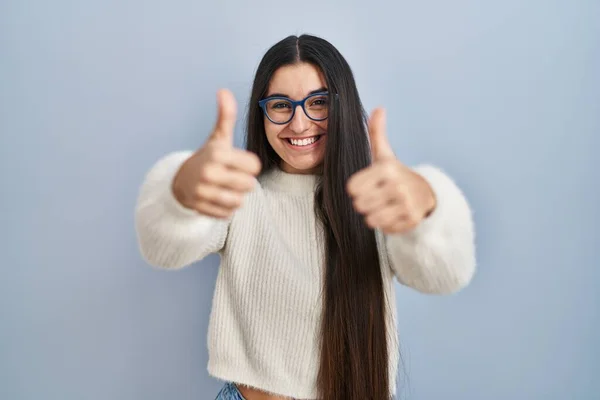 Young hispanic woman wearing casual sweater over blue background approving doing positive gesture with hand, thumbs up smiling and happy for success. winner gesture.