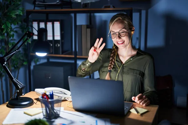 Young blonde woman working at the office at night showing and pointing up with fingers number three while smiling confident and happy.