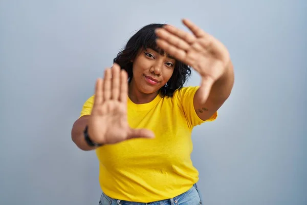Hispanic woman standing over blue background doing frame using hands palms and fingers, camera perspective