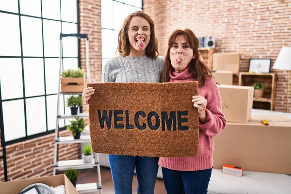 Mother and daughter holding welcome doormat sticking tongue out happy with funny expression.