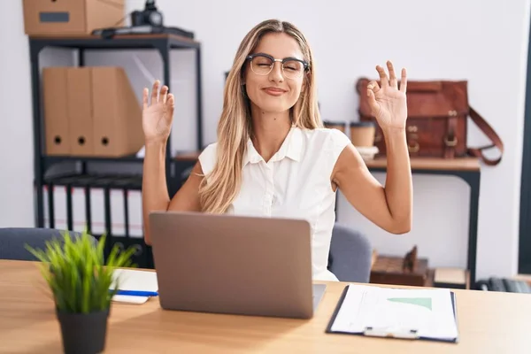 Young blonde woman working at the office wearing glasses relax and smiling with eyes closed doing meditation gesture with fingers. yoga concept.