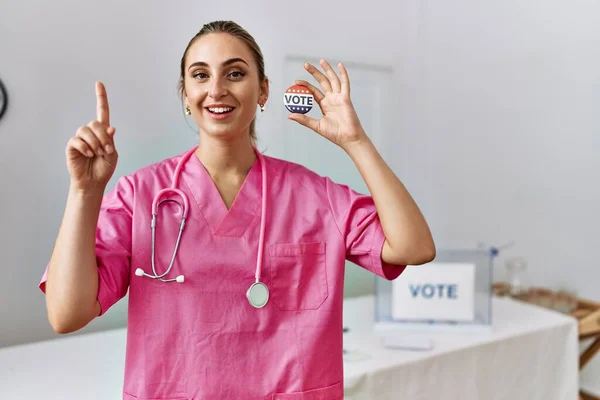 Young nurse woman at political campaign holding usa vote badge surprised with an idea or question pointing finger with happy face, number one