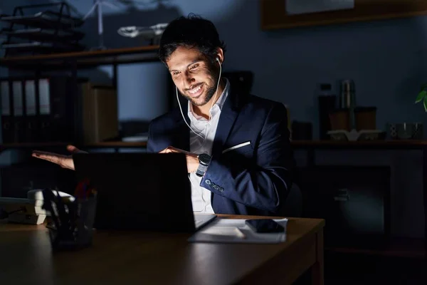 Handsome latin man working at the office at night amazed and smiling to the camera while presenting with hand and pointing with finger.