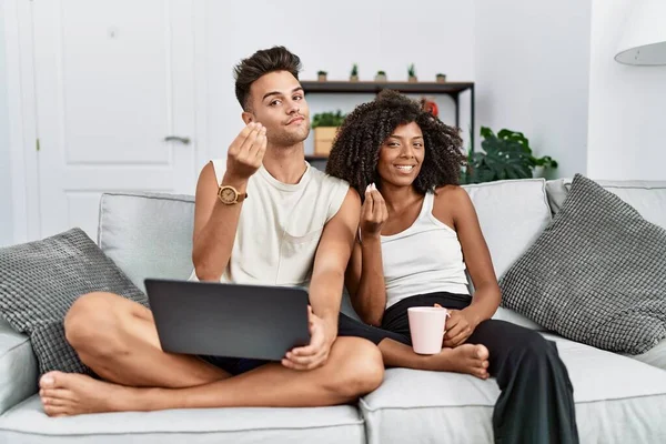 Young interracial couple using laptop at home sitting on the sofa doing money gesture with hands, asking for salary payment, millionaire business