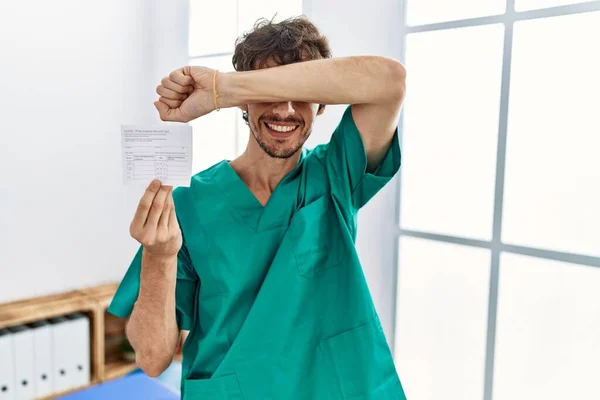 Young hispanic doctor man holding covid record card smiling cheerful playing peek a boo with hands showing face. surprised and exited