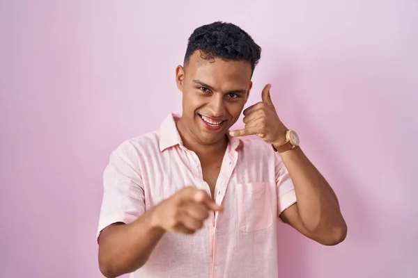 ピンクの背景の上に立っている若いヒスパニック系の男は 電話のジェスチャーで話をし あなたを指して笑顔 電話してくれ — ストック写真