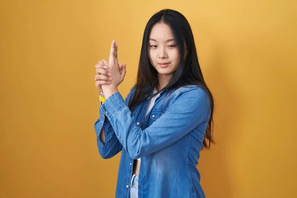 Young asian woman standing over yellow background holding symbolic gun with hand gesture, playing killing shooting weapons, angry face