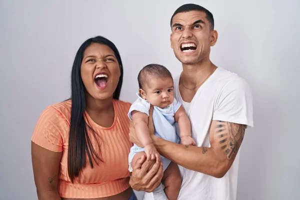 Young hispanic couple with baby standing together over isolated background angry and mad screaming frustrated and furious, shouting with anger. rage and aggressive concept.
