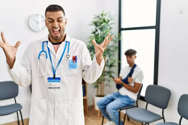 Hispanic doctor man at waiting room with pacient with arm injury celebrating mad and crazy for success with arms raised and closed eyes screaming excited. winner concept