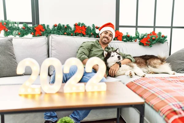 Young hispanic man hugging dog celebrate new year sitting by christmas decor at home