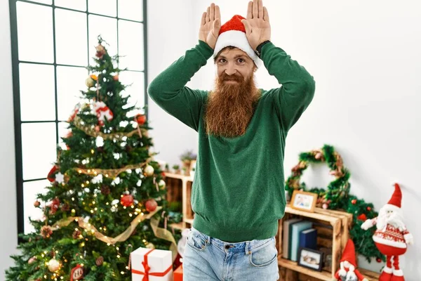 長い髭を生やした赤毛の男がクリスマスツリーでクリスマスの帽子をかぶって 手の手のひらで皮肉と懐疑的に見えるウサギの耳のジェスチャーをしています イースターウサギのコンセプト — ストック写真