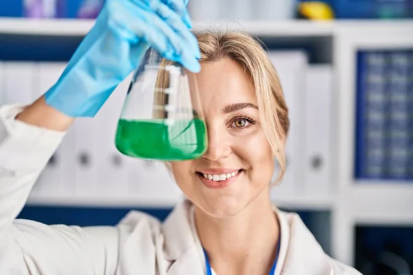 Young blonde woman scientist holding test tube over eye at laboratory