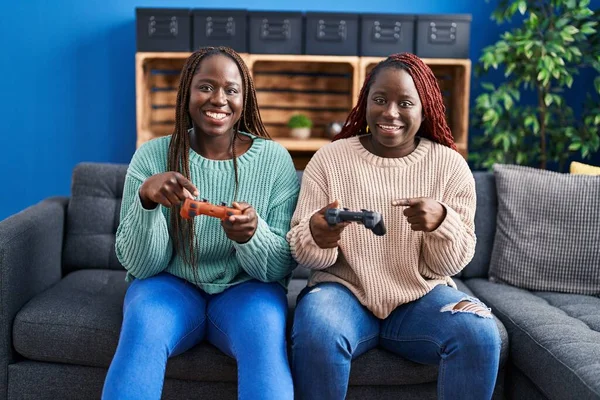 两个玩电子游戏的非洲女人牵着控制器笑着用手和手指愉快地指指点点着 — 图库照片