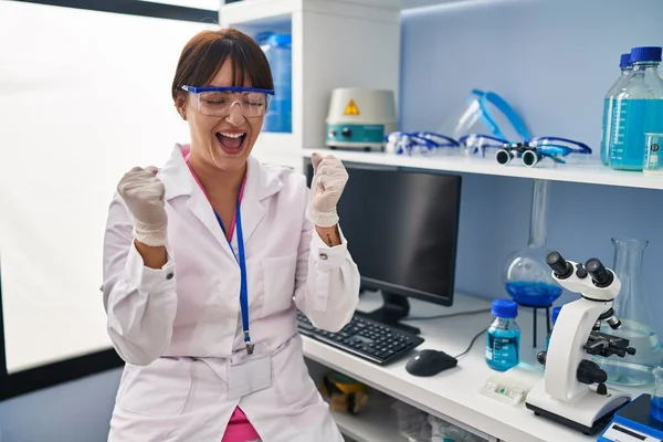 科学研究所で働く若いブルネットの女性は非常に満足し 腕を上げて勝者のジェスチャーをすることに興奮し 笑顔と成功のために叫んでいます お祝いのコンセプト — ストック写真