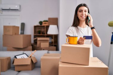 Young beautiful hispanic woman talking on smartphone holding credit card at new home
