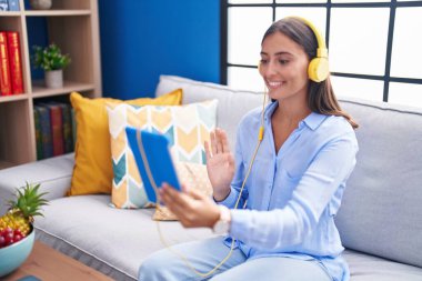 Young hispanic woman doing video call with tablet wearing headphones looking positive and happy standing and smiling with a confident smile showing teeth 