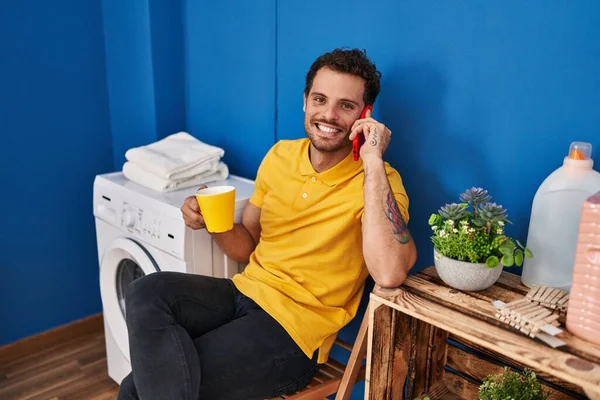Ung Latinamerikan Man Dricker Kaffe Talar Smartphone Wating För Tvättmaskin — Stockfoto