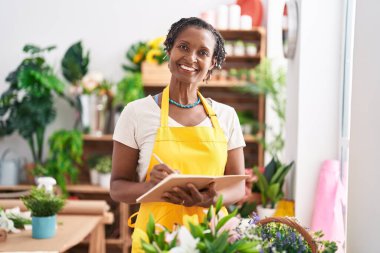 Middle age african american woman florist smiling confident writing on notebook at flower shop
