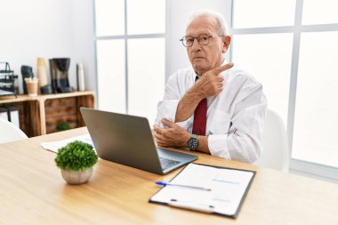 Ofiste çalışan kıdemli bir adam bilgisayarı kullanarak elini kaldırıyor ve reklamını yapıyor, ciddi ve sakin bir yüz ifadesi var. 