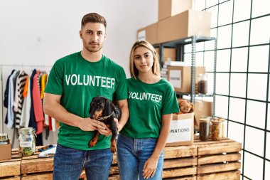 Bağışta gönüllü tişört giyen sevimli köpekli genç çift düşünceli tavır takınıyor ve kendine güvenen ayık bir ifade takınıyor. 