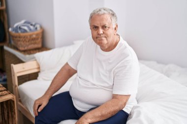 Orta yaşlı, kır saçlı, yatak odasında rahat bir ifadeyle yatakta oturan bir adam.