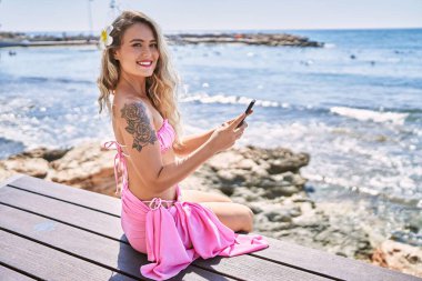 Kumsalda bankta oturan akıllı telefon kullanan genç sarışın kız..
