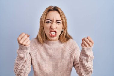 Mavi arka planda öfkeli ve kızgın duran İspanyol kadın öfkeyle bağırırken yumruklarını kaldırıyor ve sinirleniyor. öfke ve agresif konsept. 