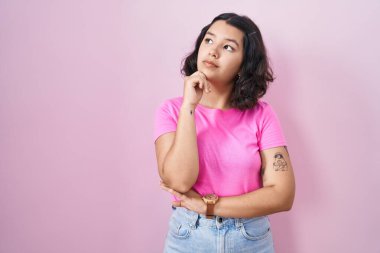 Pembe arka planda, elleri çenesinde soru soran genç İspanyol kadın, düşünceli bir ifade. Düşünceli bir yüzle gülümsüyor. şüphe kavramı. 