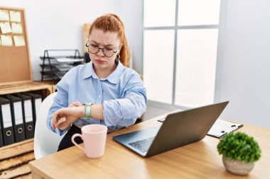 Ofiste çalışan genç kızıl saçlı bir kadın bilgisayar kullanıyor. Kol saatini kontrol ediyor, rahat ve kendinden emin. 