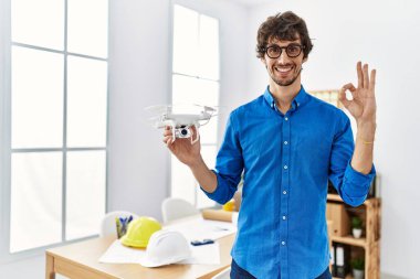 Young hispanic man using drone at architect office doing ok sign with fingers, smiling friendly gesturing excellent symbol 