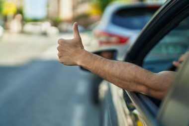 Young hispanic man driving car doing ok gesture with thumb up at street