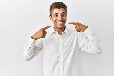 Soyutlanmış arka planda duran, neşeli gülümseyen ve parmaklarıyla ve ağzıyla işaret eden genç, yakışıklı İspanyol adam. diş sağlığı kavramı. 