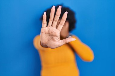 Black woman with curly hair standing over blue background covering eyes with hands and doing stop gesture with sad and fear expression. embarrassed and negative concept. 