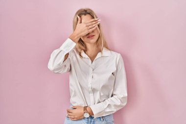 Young caucasian woman wearing casual white shirt over pink background covering eyes with hand, looking serious and sad. sightless, hiding and rejection concept 