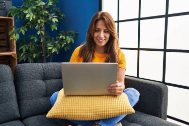 Evdeki kanepede dizüstü bilgisayar kullanan genç Latin kadın.