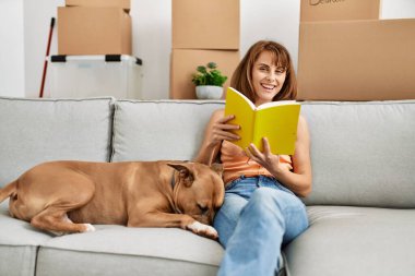 Genç beyaz kadın kitap okuyor. Kanepede köpeğiyle oturuyor.