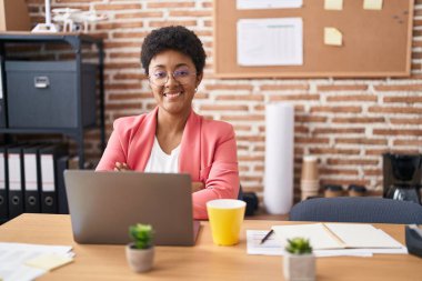 Ofiste çalışan, gözlüklü, mutlu yüzlü, kollarını kavuşturup kameraya bakan Afro-Amerikan genç bir kadın. pozitif kişi. 