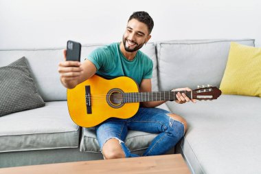 Genç Arap adam evde klasik gitar çalarak akıllı telefonun yanında selfie çekiyor.