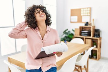 Orta Doğulu genç bir kadın iş yerinde gülümseyerek saçlarına dokunarak el hareketi yaparak çekici ve modaya uygun bir poz veriyor. 