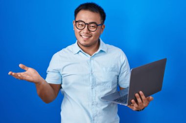 Chinese young man using computer laptop smiling cheerful presenting and pointing with palm of hand looking at the camera. 