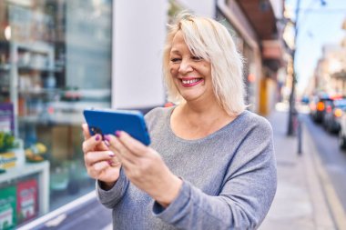 Orta yaşlı sarışın kadın sokakta akıllı telefon kullanarak gülümsüyor.