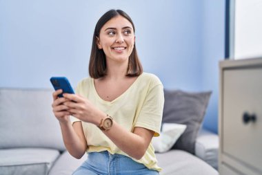 Evdeki kanepede oturan, akıllı telefon kullanan genç İspanyol kadın.