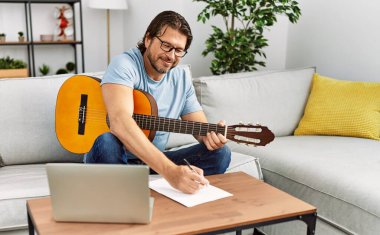 Orta yaşlı beyaz bir adam evde gitar ve dizüstü bilgisayar kullanarak şarkı besteliyor.